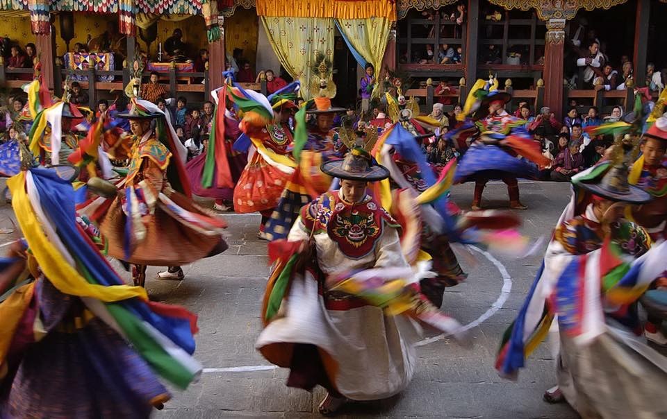 Dancers in ornate costumes and wide hats spinning coloured streamers during a festival dance in a stone dzong courtyard, spectators watching from around and above