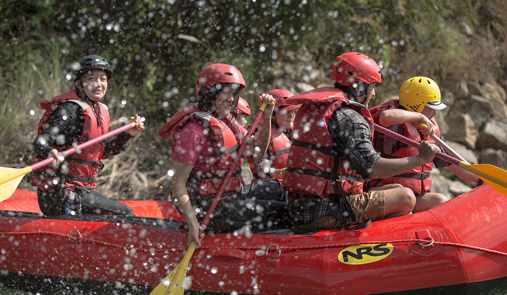 A crew in red helmets and life vests paddling a red inflatable raft through splashing white-water rapids on a Bhutanese river