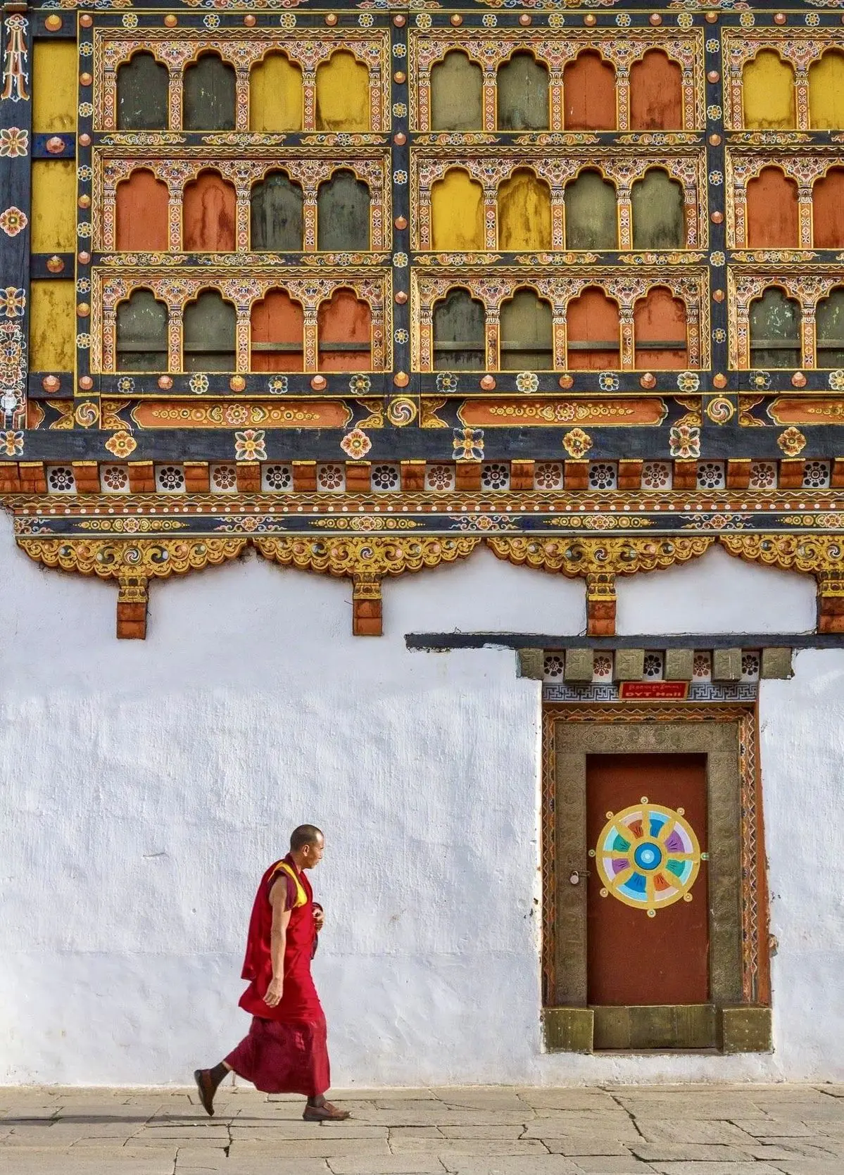 A Bhutanese monk in maroon robes passing the ornate, gold-trimmed walls of a traditional monastery