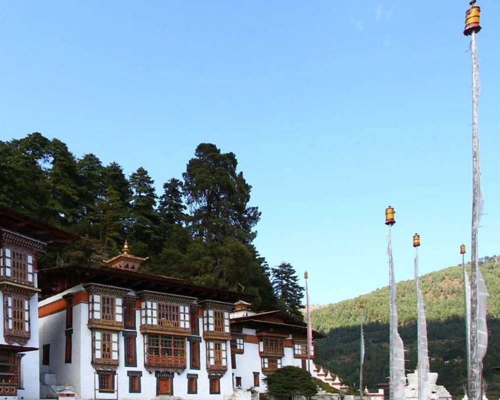 A traditional Bhutanese temple with ornate wooden windows and a golden roof, tall white prayer-flag poles raised on the grass before it